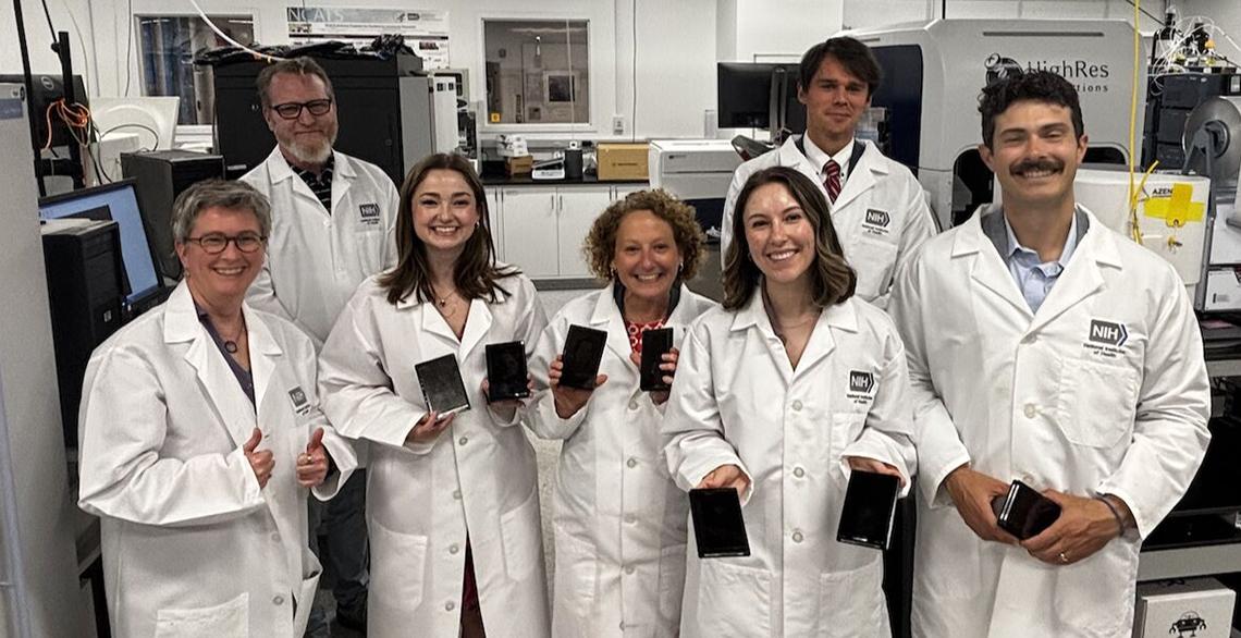 A smiling group in white lab coats in the lab hold up their micro-plates.