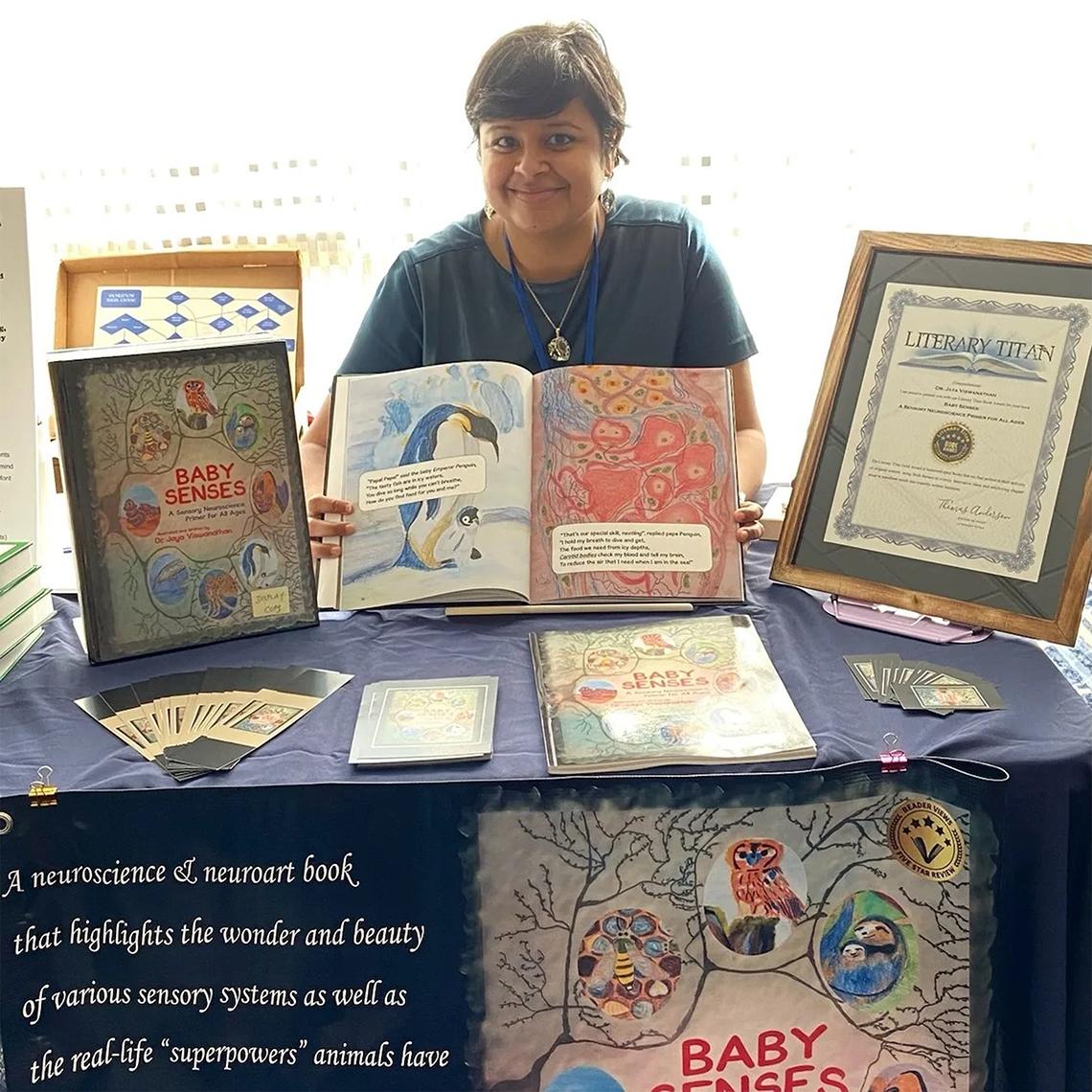 Viswanathan sits behind a desk displaying a certificate and copies of her book.