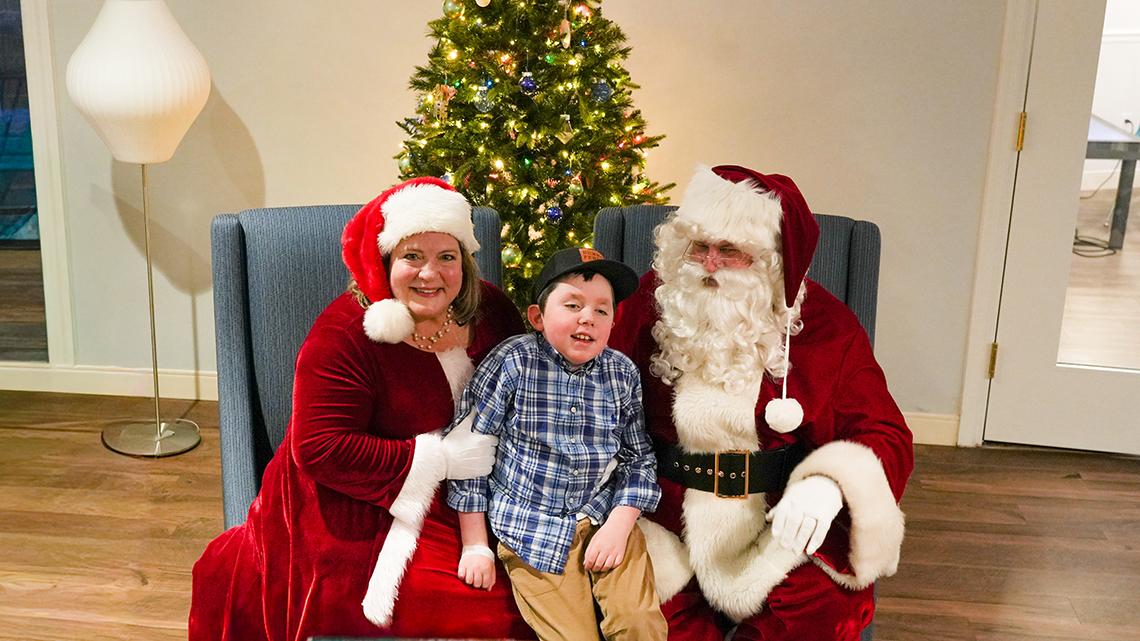 A child in a blue patterned shirt sits between Santa and Mrs. Claus on a blue chair. A Christmas tree fills the space behind the chair.